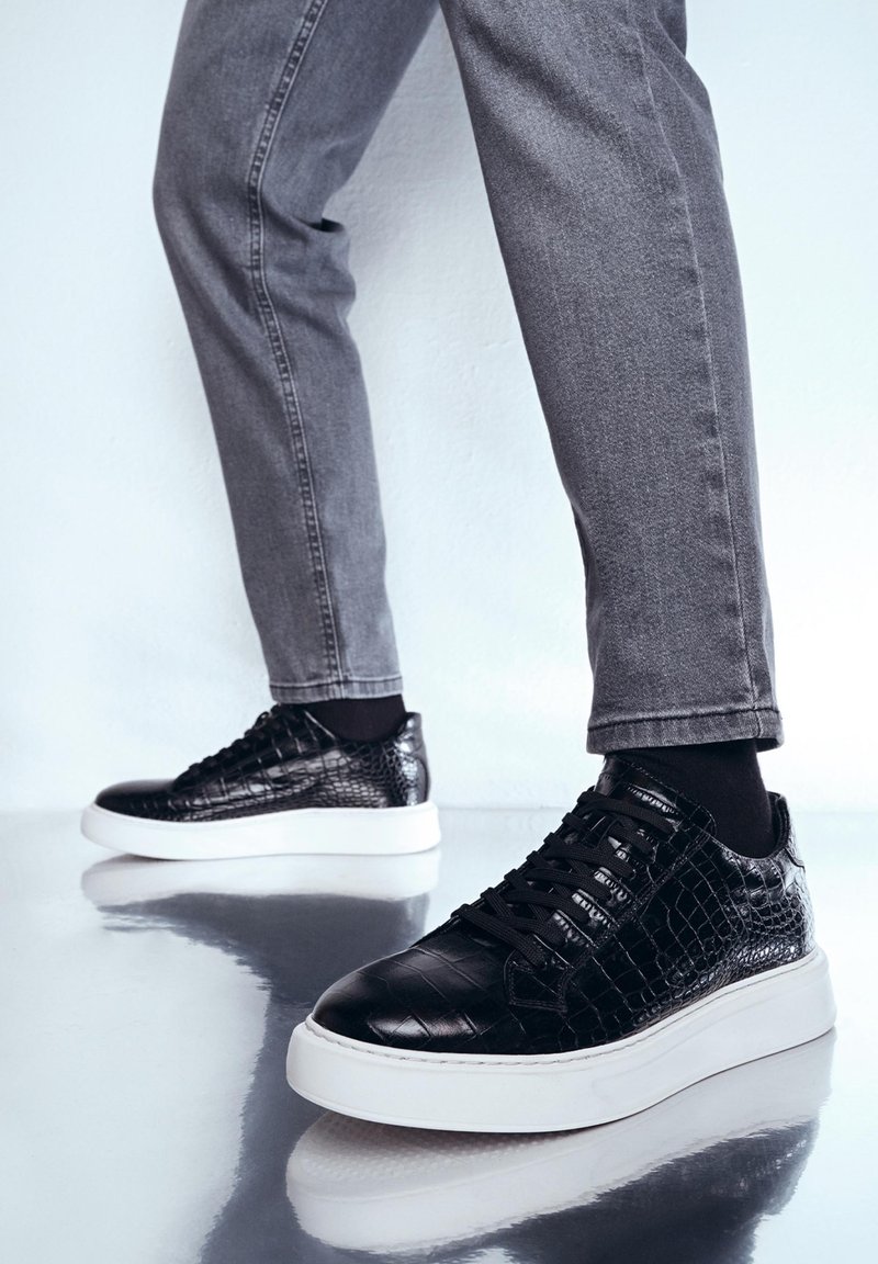 Sneaker in pelle nera con texture di coccodrillo, suola piatta bianca e lacci neri, abbinati a jeans skinny grigi.