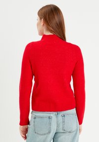 Pull en laine rouge avec un col montant, des poignets et un ourlet côtelés. Porté avec un jean en denim bleu, dévoilant un tissu texturé et une coupe ajustée.