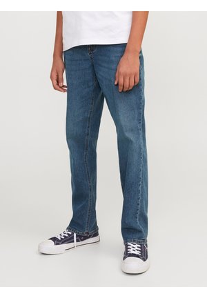 Jeune portant un jean bleu droit et des baskets bleu marine avec des lacets blancs, debout sur un fond clair uni.