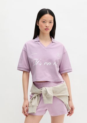 Fiatal nő levendula színű, átalakított galléros pólót visel, rajta az "It's on us" felirat, valamint hozzáillő rövidnadrágot, rajta bézs színű pulóver, amelyet a derekán kötött meg.