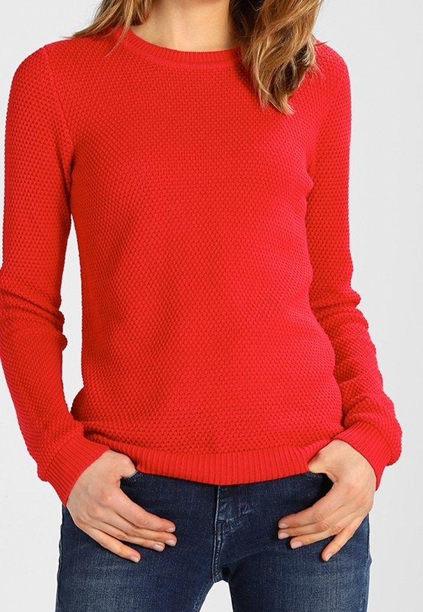 Femme portant un pull rouge texturé à manches longues et un jean bleu, debout, les mains posées sur les hanches sur un fond uni.