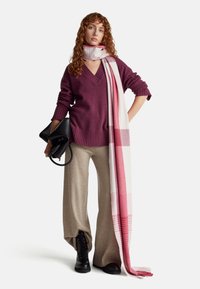 Bordeauxfarbener V-Ausschnitt-Pullover, beigefarbene weite Hose, rosa kariertes Tuch, schwarze Handtasche und schwarze Schnürstiefel. Sanfte Texturen und lockere Passform.