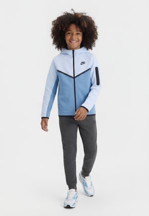 Copil zâmbitor care merge pe jos, purtând un hanorac Nike cu fermoar, albastru deschis și bleumarin, pantaloni sport gri închis și adidași albi cu albastru.