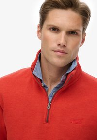 Sudadera roja con cremallera y cuello alto, de textura suave, con un logotipo bordado en el pecho, superpuesta sobre una camisa azul con cuello.