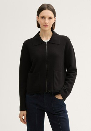 Schwarze Zip-Up-Jacke mit einem kragenförmigen Design, zwei Fronttaschen und einem strukturierten Stoff. Trägt man über einem weißen Oberteil und blauen Jeans.