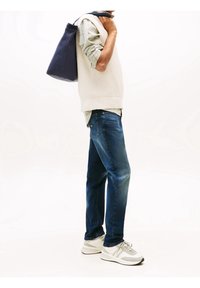 Weißer Strickweste über einem hellgrünen Hemd, blaue Jeans und grau-weiße Sneaker. Eine dunkelblaue Tasche hängt über der Schulter.