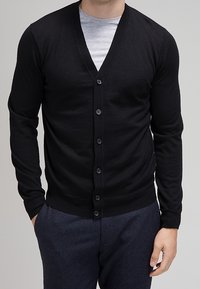 Svart cardigan med V-ringning, långa ärmar och sexknappsglidlås. Stickad struktur, bärs över en grå skjorta, kombinerad med mörka byxor.