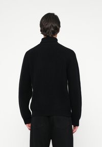 Homme aux cheveux foncés portant un pull en maille noir à col roulé et un pantalon noir, vu de dos contre un fond uni.