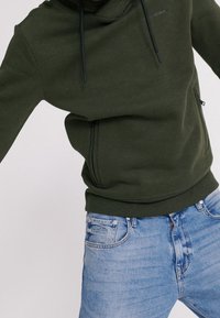 Felpa verde oliva con colletto alto, cordini e tasche laterali con zip, abbinata a jeans in denim blu chiaro con cuciture standard.