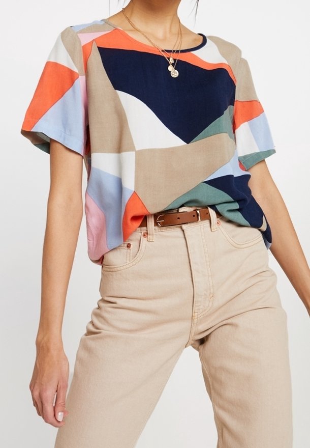 Femme portant un top à manches courtes à motif abstrait multicolore rentré dans un pantalon beige taille haute avec une ceinture marron.