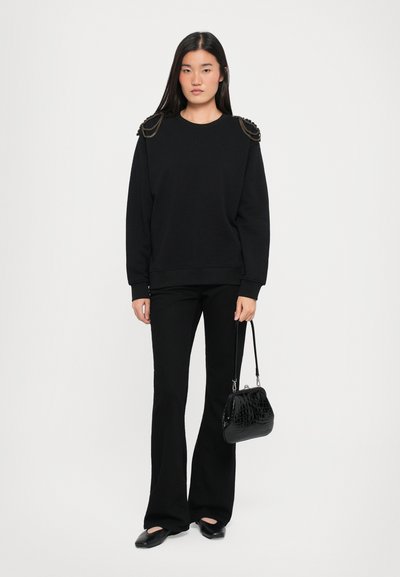 Czarna bluza z łańcuchowymi detalami na ramionach, zestawiona z czarnymi spodniami dzwonami i małą czarną torebką o fakturze. Płaskie czarne buty dopełniają całość.