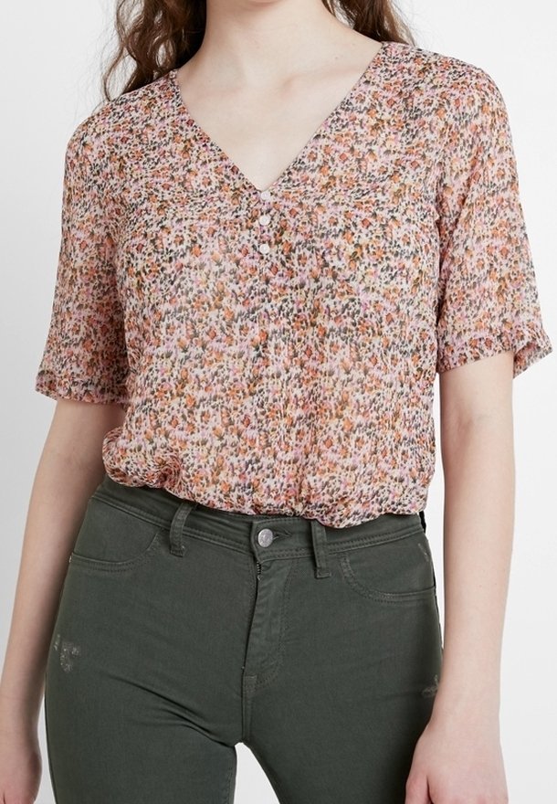 Femme portant un chemisier à manches courtes, rose à motifs floraux et col en V, rentré dans un jean taille haute vert foncé, devant un fond blanc uni.