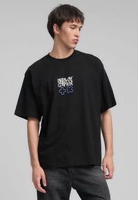T-shirt nera oversize in cotone, con una stampa grafica stilizzata bianca e blu sul davanti. Maniche corte e collo a giro.