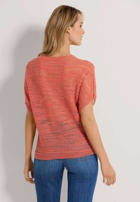 Eine Frau steht seitlich und trägt ein lockeres, korallenfarbiges, kurzärmliges Strickoberteil und blaue Jeans vor einem einfarbigen hellen Hintergrund.