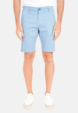 Uomo con pantaloncini azzurri fino al ginocchio, scarpe da ginnastica bianche e camicia bianca in piedi davanti a uno sfondo semplice.