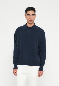 Uomo che indossa un maglione a polo a maniche lunghe blu navy e pantaloni bianchi, in piedi contro uno sfondo bianco semplice.