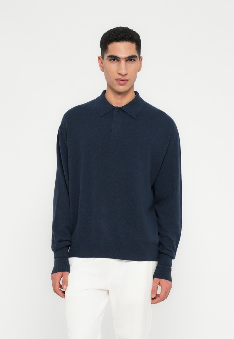 Uomo che indossa un maglione a polo a maniche lunghe blu navy e pantaloni bianchi, in piedi contro uno sfondo bianco semplice.