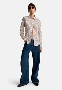 Camicetta a fiori con bottoni, con motivi rosa e verdi, abbinata a jeans larghi blu scuro e scarpe nere a punta. Inclusi occhiali da sole.