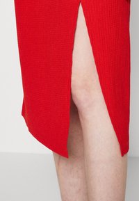 Robe tricotée rouge avec une texture côtelée et une fente sur le côté, révélant la peau en dessous. L'ourlet est inégal, ajoutant un détail dynamique.