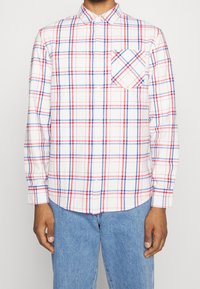 Man i en vit skjorta med knappar i ett rutmönster av röd och blå färg, med en bröstficka, ihop med ljusblå jeans.