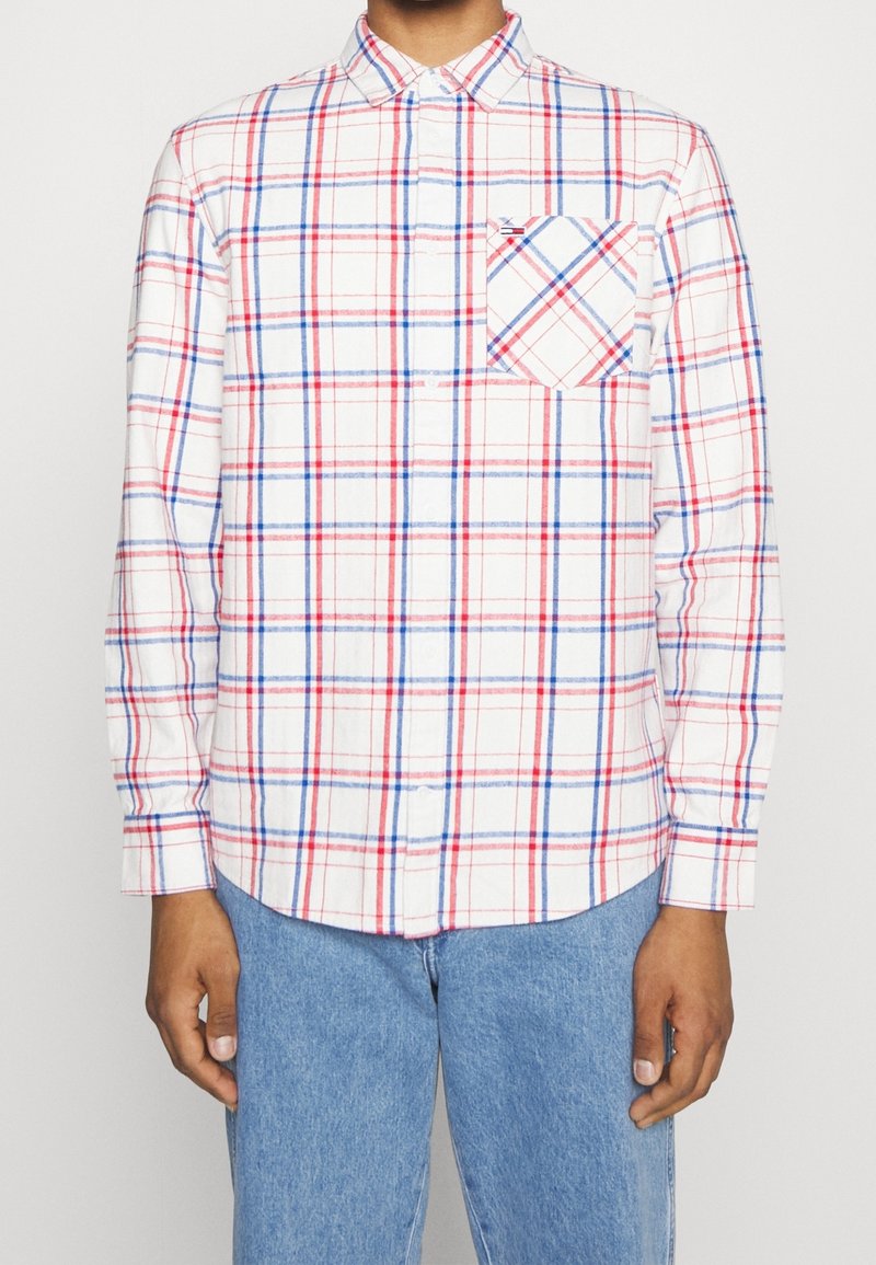 Man i en vit skjorta med knappar i ett rutmönster av röd och blå färg, med en bröstficka, ihop med ljusblå jeans.