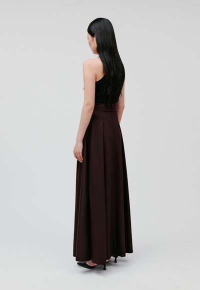 Maxi skirt in dark burgundy fabric, features pleats, flared silhouette, high waistband, paired with a black sleeveless top and heels.