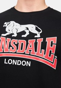 Zwarte katoenen t-shirt met een witte leeuwafbeelding en vet rood "Lonsdale"-tekst met "London" eronder in het wit. Zachte textuur, ronde hals ontwerp.