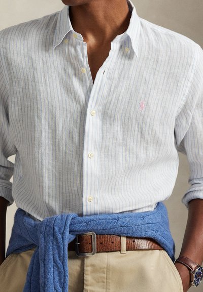 Λευκό πουκάμισο από λινό με ανοιχτό μπλε και λευκές ρίγες, με γιακά με κουμπιά, διακοσμημένο με μικρό ροζ λογότυπο και μανίκια σηκωμένα. Μπλε πλεκτό πουλόβερ δεμένο γύρω από τη μέση.