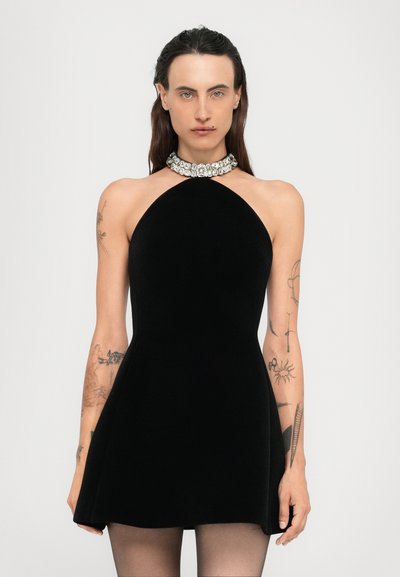 Femme aux longs cheveux noirs portant une robe noire sans manches avec un col en pierres précieuses, montrant des tatouages sur les deux bras, se tenant devant un fond uni.