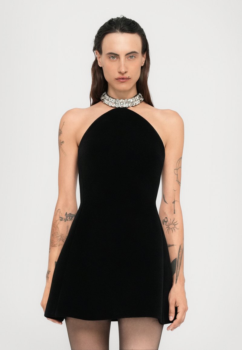 Kvinna med långt mörkt hår, klädd i en ärmlös svart klänning med ett smyckat krage, med tatueringar på båda armarna, stående mot en enfärgad bakgrund.