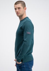 Bawełniana bluzka dresowa w kolorze morskim z okrągłym dekoltem, ściągaczami na rękawach i dole, z naszywką na lewym rękawie. Dopasowany fason, widok z boku.