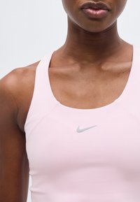 Κοντινό πλάνο ενός ατόμου που φορεί ανοιχτό ροζ αθλητικό σουτιέν Nike, δείχνοντας το λαιμό, τους ώμους και το πάνω μέρος του στήθους με ορατό λογότυπο.