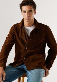 Καφέ σατέν μπουφάν με κουμπιά στο μπροστινό μέρος, μία τσέπη στο στήθος και ασημί μεταλλικά στοιχεία. Συνδυασμένο με ανοιχτό μπλε τζιν παντελόνι.