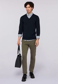 Marineblauer Pullover mit V-Ausschnitt über einem blau-weiß gestreiften Hemd, olivgrüne Hose, schwarze Schuhe und eine schwarze Einkaufstasche. Glattes, eng anliegendes Design.