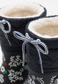 Bottes d'hiver matelassées bleu foncé avec des motifs floraux blancs, doublure en fausse fourrure beige douce, et lacets tressés blancs et bleu marine contrastants.