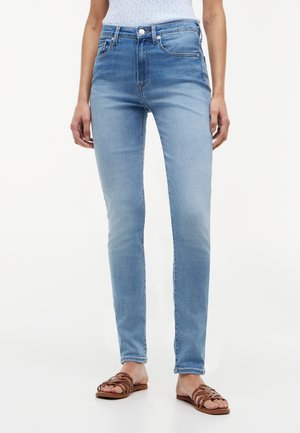 Kvinde iført lyseblå slim-fit jeans og brune sandaler med åbne tæer, stående mod en hvid baggrund.