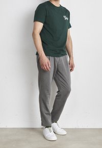 Tmavě zelené tričko s bílým logem, šedé zúžené kalhoty a bílé tenisky. Hladký materiál, volný střih, minimalistický design a kontrastní barvy.