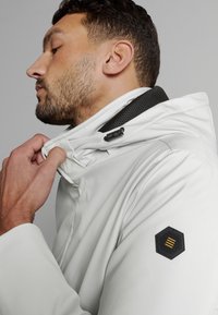Witte waterdichte jas met een hoge kraag, voorzien van een zwart gaasaccent, zijrits en een hexagonaal logo-opdruk met gele strepen.