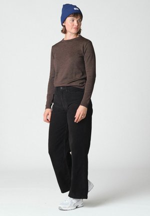 Jeune personne portant une chemise marron à manches longues, un pantalon noir à jambes larges, un bonnet tricoté bleu et des baskets blanches, debout devant un fond uni.