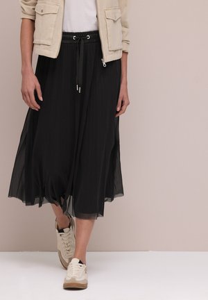 Femme portant une veste beige, un haut blanc, une jupe midi plissée noire avec taille à cordon, et des baskets crème, debout devant un fond uni.