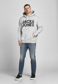 Grijze hoodie met groot zwart "JACK & JONES" logo, gecombineerd met een spijkerbroek en zwarte sneakers. Model staat tegen een neutrale achtergrond.