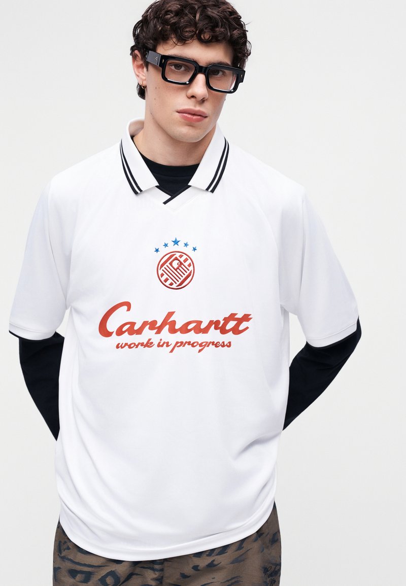 Junger Mann mit lockigem Haar und großen schwarzen Brillen, der ein weißes Carhartt-Poloshirt über einem schwarzen Langarmshirt und gemusterten Hosen trägt.