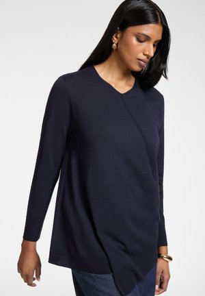 Vrouw met lang zwart haar, gekleed in een marineblauw asymmetrisch shirt met lange mouwen, gouden oorringen en een gouden armband, kijkt naar beneden.