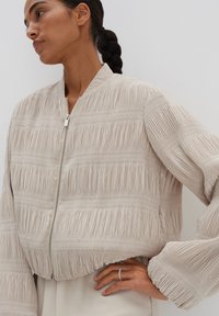 Helles beiges Bomberjacke mit plissierter Textur, Reißverschluss vorne und gerippten Bündchen. Der Stoff hat einen sanften Glanz und ein horizontales Streifenmuster.