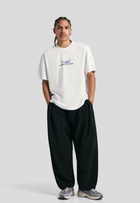 White t-shirt with purple text, oversized fit. Black baggy trousers. Feet in gray sneakers with red accents. Simple, casual streetwear style.