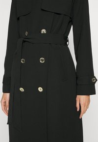Trench-coat noir croisé avec boutons dorés, ceinture à la taille nouée, manches longues avec poignets boutonnés, porté sur le torse et les bras d'une personne.