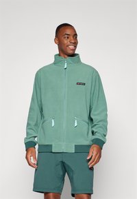 Grønn fleece zip-up jakke med høy krage, to glidelåslommer og ribbestrikkede ermer; brukt med matchende grønne shorts. Merkevarelogo på brystet.