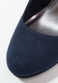 Gros plan sur la pointe d’une chaussure en daim bleu marine à forme arrondie avec des coutures visibles le long du bord sur un fond clair.