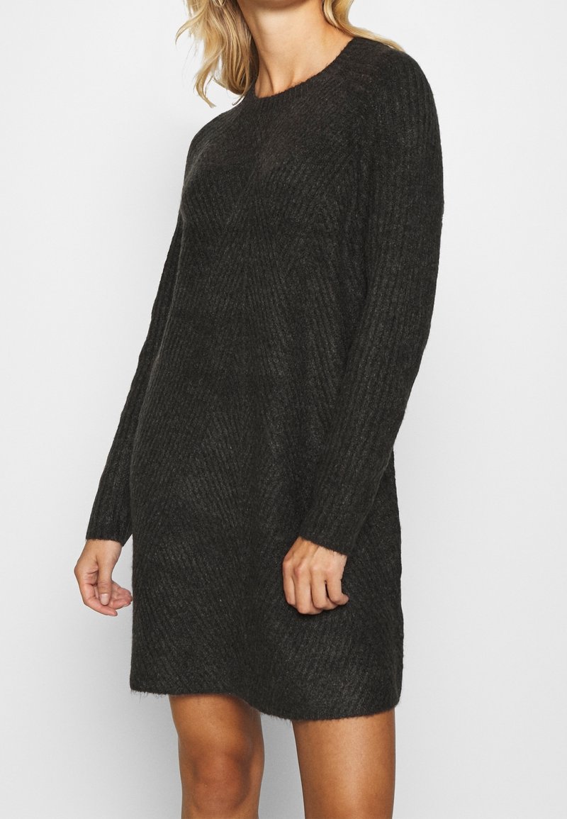 Femme portant une robe-pull en maille côtelée gris foncé à manches longues, debout devant un fond blanc uni.