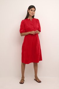 Rochie scurtă roșie cu mâneci scurte, guler, detalii cu nasturi și o croială lejeră, realizată dintr-un material neted. Poartă sandale plate.